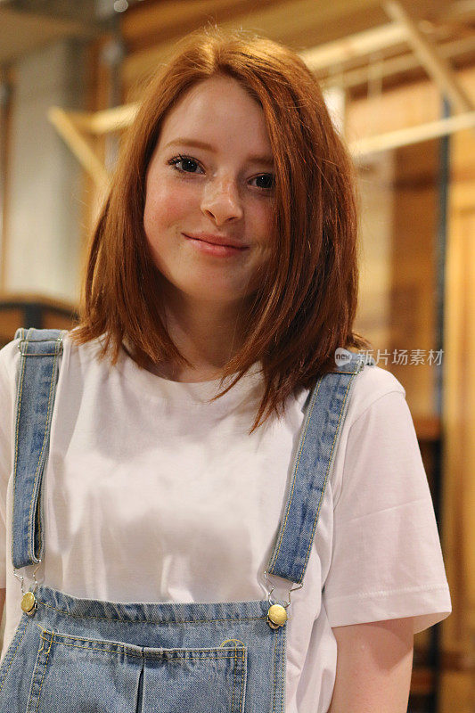 14 / 15岁的红发少女站在商店里，穿着休闲的蓝色牛仔工装裤，外面是一件朴素的白色t恤